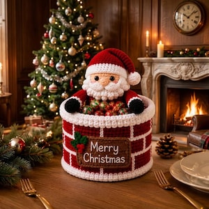 Può includere: Una figura di Babbo Natale all'uncinetto che emerge da un cesto con motivo a mattoni rossi e bianchi. Il cesto presenta una toppa rettangolare marrone con il testo Merry Christmas e foglie di agrifoglio verdi. È riempito di ornamenti rotondi.