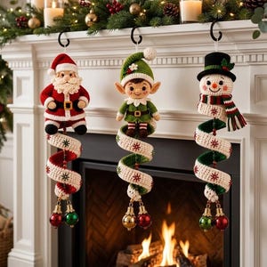 May include: Three crochet Christmas ornaments hanging from black hooks. The left is a Santa Claus, the center is an elf, and the right is a snowman. Each figure is attached to a long, spiraling, cream-colored fabric tail with red and green accents.