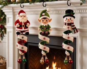 May include: Three crochet Christmas ornaments hanging from black hooks. The left is a Santa Claus, the center is an elf, and the right is a snowman. Each figure is attached to a long, spiraling, cream-colored fabric tail with red and green accents.