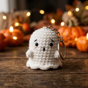 Puede incluir: Un pequeño llavero de fantasma tejido a ganchillo en color blanco, con cabeza redondeada, dos brazos pequeños y un rostro con ojos negros bordados, una boca circular pequeña y mejillas rosadas, unido a una anilla de metal plateado.