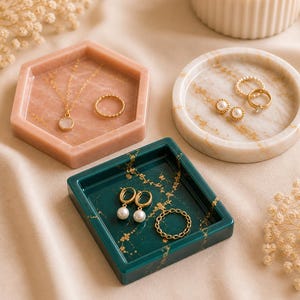 May include: A collection of jewelry trays in various shapes and colors. A hexagonal pink tray holds a gold necklace and ring. A round marble tray displays earrings and rings. A square teal tray holds earrings and a ring. Gold accents are visible.