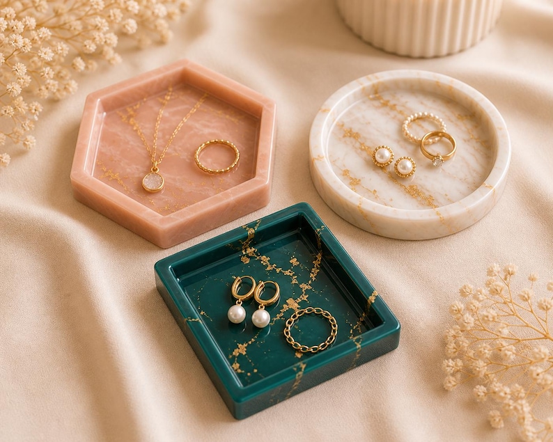 May include: A collection of jewelry trays in various shapes and colors. A hexagonal pink tray holds a gold necklace and ring. A round marble tray displays earrings and rings. A square teal tray holds earrings and a ring. Gold accents are visible.