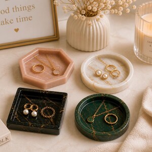May include: A collection of jewelry trays in various shapes and colors, including pink, white, black, and green, each holding rings and necklaces. A lit candle and a framed print with the text "good things take time" are also present.