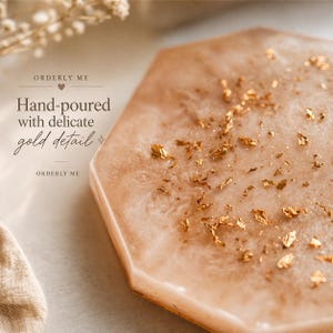 May include: A hexagonal, hand-poured coaster with a pearlescent, light peach hue. The coaster is adorned with delicate gold leaf details. The text "ORDERLY ME" and "Hand-poured with delicate gold detail" are visible.