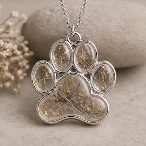 May include: A silver paw print pendant filled with clear resin, containing dark hair and gold flakes. The pendant hangs from a delicate silver chain. A memorial keepsake, the pendant is a touching tribute.