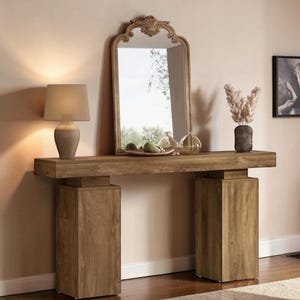 May include: A wooden console table with a rectangular top and two square supports. A decorative mirror with an ornate top is centered on the table. A lamp and vase with dried flowers are on the table. The room has a neutral color palette.