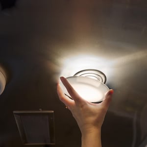 May include: A round, frosted glass light fixture is held in place. The fixture is mounted on a metallic surface, with a bright light emanating from the center. A circular speaker and a rectangular vent are also visible.