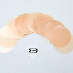 Puede incluir: Una colección de cubiertas de pezones circulares translúcidas de color piel dispuestas en arco. Una púa blanca con el texto "AirShade" en un rectángulo negro está en la parte inferior.
