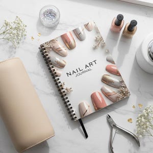 Op de afbeelding: Een Nail Art Journal met spiraalbinding, met nagelontwerpen in roze, goud en wit. De afbeelding bevat nagellakflesjes, nagelknippers, een beige etui en een kleine container met nail art versieringen. De kaft van het tijdschrift luidt "NAIL ART JOURNAL."