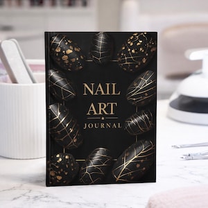 Op de afbeelding: Een zwart "NAIL ART JOURNAL" met gouden details. De omslag toont zwarte nagelontwerpen met gouden aderen en spikkels. Het tijdschrift ligt op een wit aanrecht.