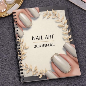 Könnte beinhalten: Ein spiralgebundenes "NAIL ART JOURNAL" Notizbuch mit beigem Einband, verziert mit einem goldenen Blattdesign und Illustrationen von manikürten Nägeln. Das Journal liegt auf einer dunkelgrauen Oberfläche, mit Nailart-Zubehör im Hintergrund.