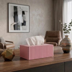 Puede incluir: Un dispensador de pañuelos rectangular rosa con líneas verticales, que contiene pañuelos blancos. El dispensador está sobre una superficie de madera oscura, junto a objetos geométricos dorados y un jarrón con vegetación. La habitación tiene una paleta de colores neutros.