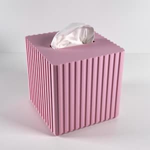 Puede incluir: Un dispensador de pañuelos cuadrado de color rosa claro con detalles verticales acanalados. La parte superior tiene una abertura cuadrada para dispensar pañuelos. Los pañuelos blancos son parcialmente visibles.