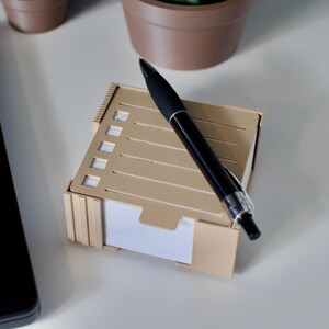 Op de afbeelding: Een kleine, beige notitieblok met een checklist ontwerp en een zwarte pen. Het notitieblok is rechthoekig met uitgesneden vakjes om items af te vinken. De pen is glanzend zwart met zilveren accenten. Op een witte ondergrond.