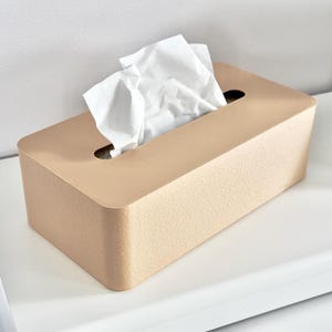 May include: A light tan rectangular tissue box with a textured finish. White tissues protrude from the oval opening on top. The box is placed on a white surface.