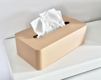 Könnte beinhalten: Eine rechteckige Taschentuchbox in hellem Beige mit strukturierter Oberfläche. Weiße Taschentücher ragen aus der ovalen Öffnung oben heraus. Die Box steht auf einer weißen Oberfläche.