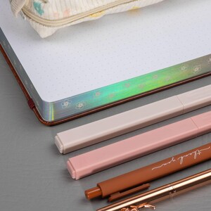 May include: A cream-colored pencil case with a floral pattern rests on a dotted notebook with a brown cover. Several pens in shades of pink, beige, and brown are arranged below, one with the words "thank you" and another with a floral design.