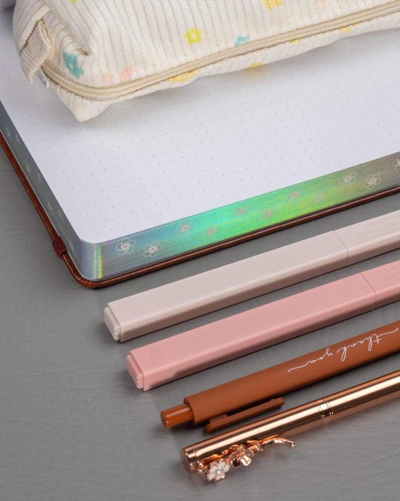 May include: A cream-colored pencil case with a floral pattern rests on a dotted notebook with a brown cover. Several pens in shades of pink, beige, and brown are arranged below, one with the words "thank you" and another with a floral design.