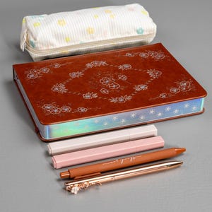 May include: A brown leather journal with floral designs, a pencil case, and several pens. The pens are in shades of pink, brown, and rose gold. One pen has the words "thank you" on it.