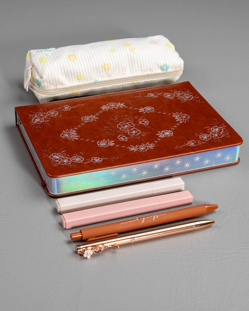 May include: A brown leather journal with floral designs, a pencil case, and several pens. The pens are in shades of pink, brown, and rose gold. One pen has the words "thank you" on it.
