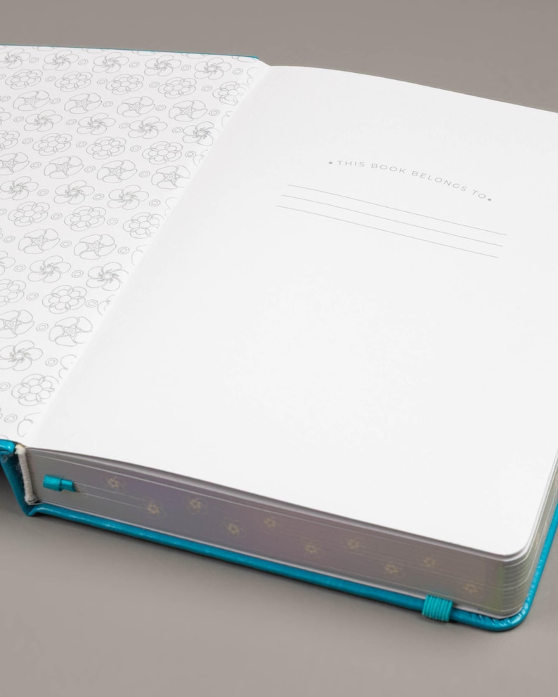 May include: Open journal with a turquoise cover and a matching ribbon bookmark. The inside pages are white, with a floral pattern on the left page and lines for writing on the right. The text "THIS BOOK BELONGS TO" is printed at the top of the right page.