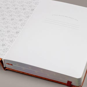 May include: Open notebook with a copper-colored cover and a red ribbon bookmark. The inside pages are white, with a floral pattern on the left page and lines for writing on the right. The text "THIS BOOK BELONGS TO." is printed at the top of the right page.