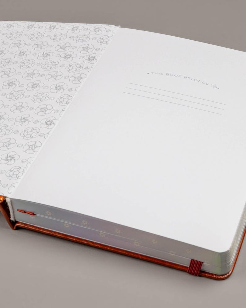 May include: Open notebook with a copper-colored cover and a red ribbon bookmark. The inside pages are white, with a floral pattern on the left page and lines for writing on the right. The text "THIS BOOK BELONGS TO." is printed at the top of the right page.