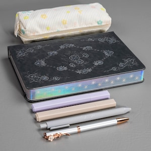 May include: A stationery set featuring a black journal with floral designs, a pencil case with a floral pattern, and several pens. The pens are in shades of gray, beige, and lavender, with a decorative floral clip on one pen.