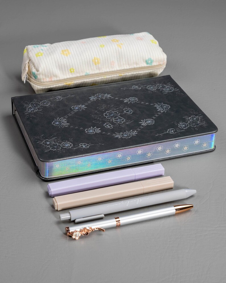 May include: A stationery set featuring a black journal with floral designs, a pencil case with a floral pattern, and several pens. The pens are in shades of gray, beige, and lavender, with a decorative floral clip on one pen.