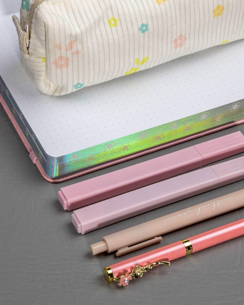 May include: A stationery set featuring a floral-patterned pencil case, a dot grid notebook, and several pink pens. One pen has the words "thank you" printed on it, and another has a decorative floral design.