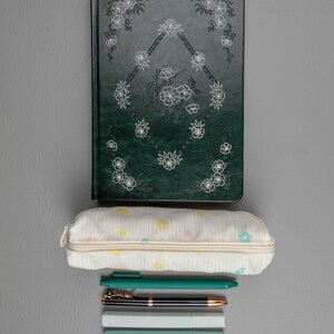 May include: A dark green journal with floral designs and a matching pen, a pencil case with pastel floral patterns, and several pens and highlighters on a gray surface. Stationery supplies for school or office.