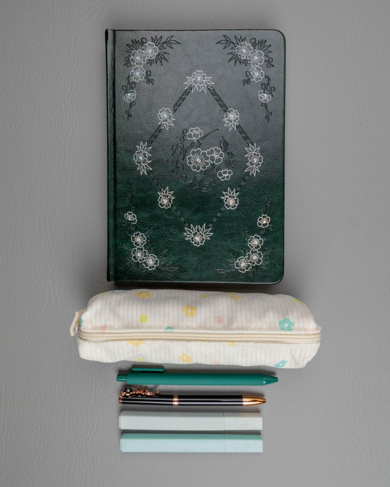 May include: A dark green journal with floral designs and a matching pen, a pencil case with pastel floral patterns, and several pens and highlighters on a gray surface. Stationery supplies for school or office.