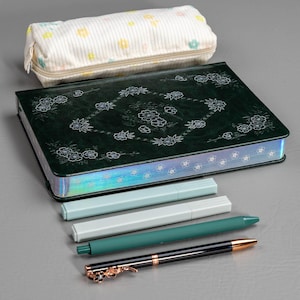 May include: A stationery set featuring a dark green journal with floral designs, a pencil case, and several pens. The pens are in shades of green, white, and black, with gold accents. The pencil case is white with a floral pattern.