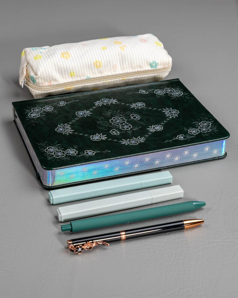 May include: A stationery set featuring a dark green journal with floral designs, a pencil case, and several pens. The pens are in shades of green, white, and black, with gold accents. The pencil case is white with a floral pattern.