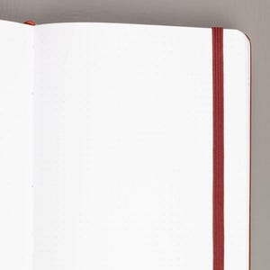 May include: Open notebook with dot grid pages and a red elastic band closure. The notebook has a brown cover and a bookmark. The pages are white with small gray dots, perfect for journaling or note-taking.
