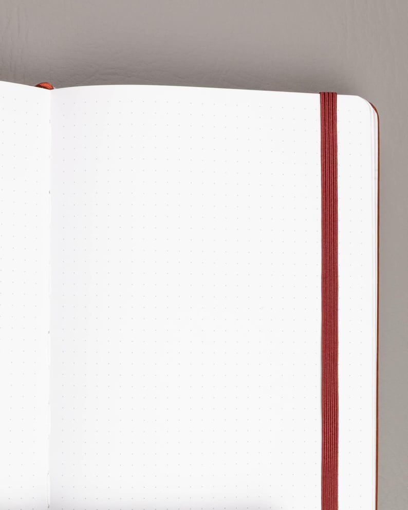 May include: Open notebook with dot grid pages and a red elastic band closure. The notebook has a brown cover and a bookmark. The pages are white with small gray dots, perfect for journaling or note-taking.