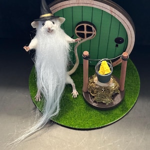 May include: A whimsical miniature scene featuring a white mouse dressed as a wizard with a long white beard and a black hat. The mouse stands before a green hobbit door. A small well with a bucket of cheese sits nearby, all on a green circular base.