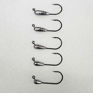 May include: Five fishing jig heads with silver-colored weights and black hooks are arranged vertically against a white background. The hooks are curved and have a small loop at the top for attaching to fishing line.