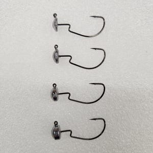 May include: Four black fishing jig heads with silver-colored weights. Each jig head has a curved hook extending from the weight. The hooks are arranged vertically against a light background.