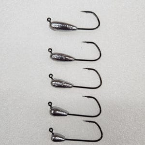 May include: Five silver-colored fishing jigs with black hooks are arranged vertically against a white background. Each jig has a teardrop-shaped head and a curved hook. The hooks are pointed and ready for use.