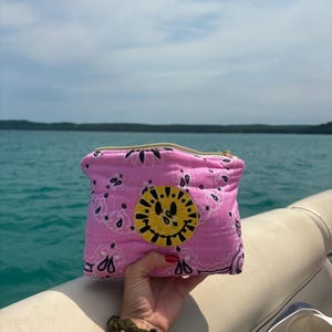 Puede incluir: Un estuche rosa con cremallera que presenta un patrón de cachemira estilo bandana en blanco y negro y un parche circular amarillo con un diseño de cara sonriente bordado en negro en el centro del accesorio de tela.