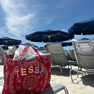 May include: A red bandana-print tote bag with the name "TESSA" in colorful letters. The bag is filled with white items and sits on a beach chair. Blue umbrellas and lounge chairs are in the background under a bright blue sky.