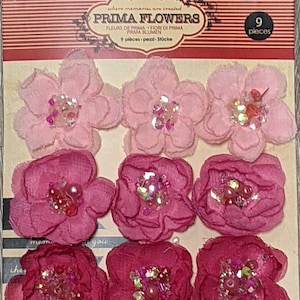 May include: Nine pink and red fabric flowers with glitter and pearl embellishments. The flowers are arranged in three rows on a brown card with the text "Prima Flowers" and "9 pieces".