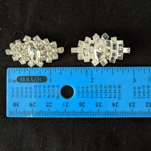 May include: Two silver-toned rhinestone brooches. One brooch features a central oval rhinestone surrounded by smaller stones. The other brooch has a similar design. A blue ruler is placed next to the brooches.