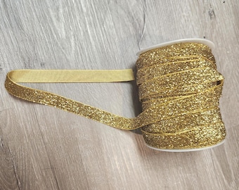 May include: A spool of gold glitter ribbon. The ribbon is unwound, revealing a flat, shimmering surface. The spool is white, and the ribbon is a bright, festive gold color. The ribbon is likely used for crafting, gift wrapping, or other decorative purposes.