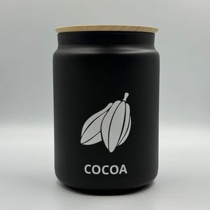 Könnte beinhalten: Ein schwarzer Kakaobehälter mit hellem Holzdeckel. Der Behälter hat eine weiße Grafik von Kakaoschoten und das Wort "COCOA" in weißer Schrift. Der Behälter ist zylindrisch und steht auf einer weißen Oberfläche.