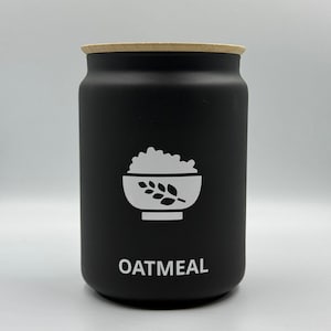 Könnte beinhalten: Ein schwarzer zylindrischer Behälter mit einem hellen Holzdeckel. Der Behälter hat eine weiße Grafik einer Schüssel mit Haferflocken und das Wort "OATMEAL" in weißer Schrift. Das Design ist schlicht und modern.
