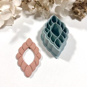 Op de afbeelding: Een lichtroze, druppelvormige oorbel en een turquoise oorbeluitsteker liggen op een wit oppervlak, naast witte en beige gedroogde bloemen. De oorbel heeft een gelaagd ontwerp en de uitsteker heeft een gedetailleerd, geometrisch patroon.