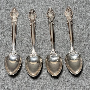May include: Four silver spoons with ornate handles and oval bowls are arranged on a gray textured surface. The spoons have a decorative floral design at the top of the handle. The spoons are reflective and appear to be made of metal.