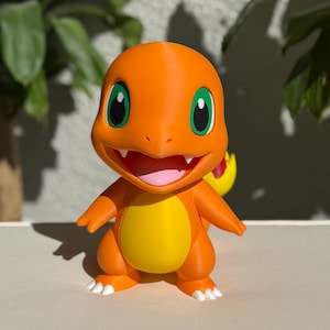 Könnte beinhalten: Orangefarbene Charmander-Figur mit grünen Augen, gelbem Bauch und einer Flamme am Schwanz. Die Figur hat einen lächelnden Ausdruck und steht auf einer weißen Oberfläche.